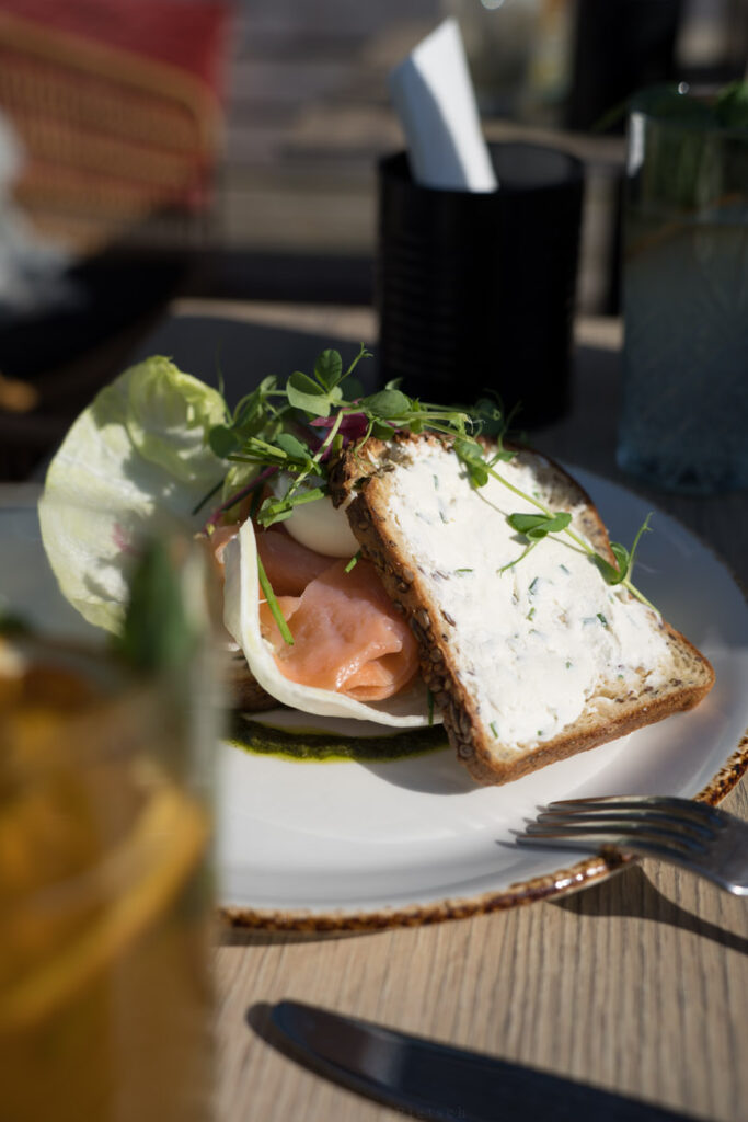 Ein Teller mit einem Brot steht auf einem Tisch in einem Café. Das Brot ist mit Frischkäse bestrichen und mit geräuchertem Lachs belegt.