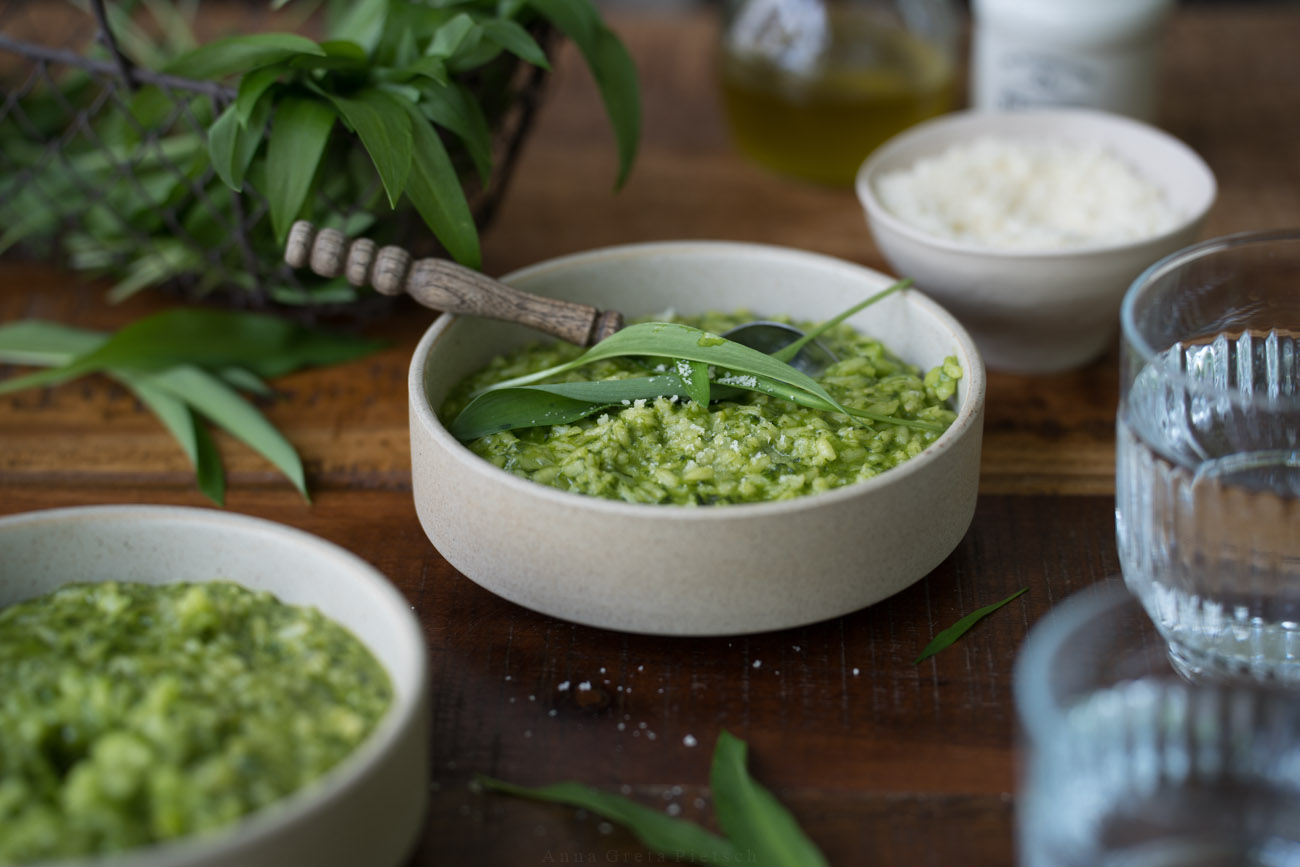 Ein Schälchen mit grünem Bärlauch-Risotto steht in der Mitte auf einer dunken Holztischplatte. Im Hintergrund sieht man ein kleines Körbchen aus draht mit Bärlauch. Vorne sieht man im Anschnitt ein Wasserglas und eine weitere Schale mit Risotto.