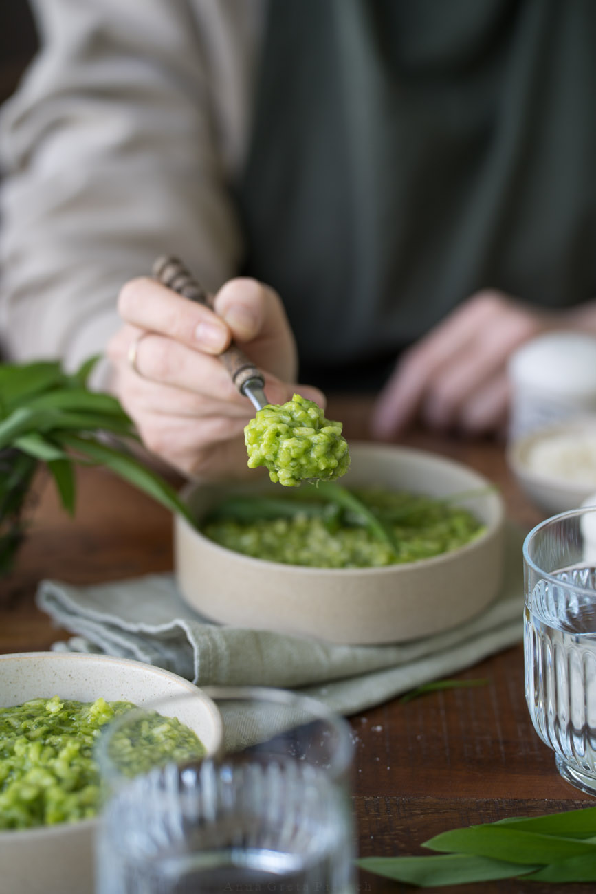 Ein Schälchen mit grünem Bärlauch-Risotto steht in der Mitte auf einer dunken Holztischplatte. Im Hintergrund sieht man ein kleines Körbchen aus draht mit Bärlauch. Vorne sieht man im Anschnitt ein Wasserglas und eine weitere Schale mit Risotto. Eien Person hält einen Löffel mit Risotto in die Mitte des Bildes/in die Kamera.