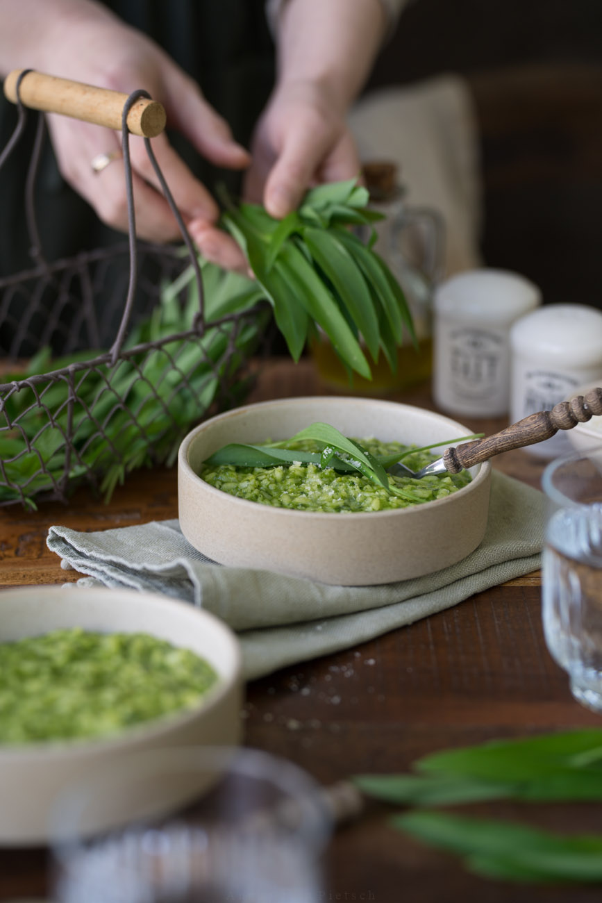 Ein Schälchen mit grünem Bärlauch-Risotto steht in der Mitte auf einer dunken Holztischplatte. Im Hintergrund sieht man ein kleines Körbchen aus draht mit Bärlauch. Vorne sieht man im Anschnitt ein Wasserglas und eine weitere Schale mit Risotto. Eine Person hält etwas frischen Bärlauch im Hintergrund in ihren Händen.