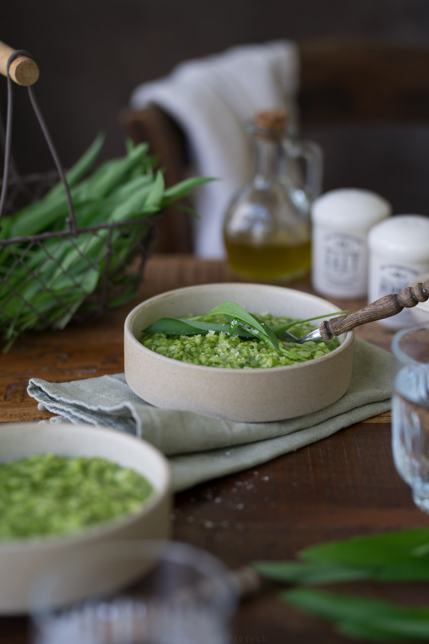 Ein Schälchen mit grünem Bärlauch-Risotto steht in der Mitte auf einer dunken Holztischplatte. Im Hintergrund sieht man ein kleines Körbchen aus draht mit Bärlauch. Vorne sieht man im Anschnitt ein Wasserglas und eine weitere Schale mit Risotto.