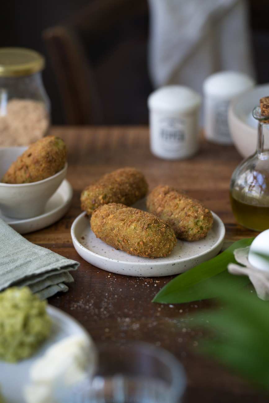 Ein Tellerchen mit drei frittierten Bärlauch-Suppli steht in der Mitte auf einer dunken Holztischplatte. Vorne sieht man im Anschnitt frischen Bärlauch.