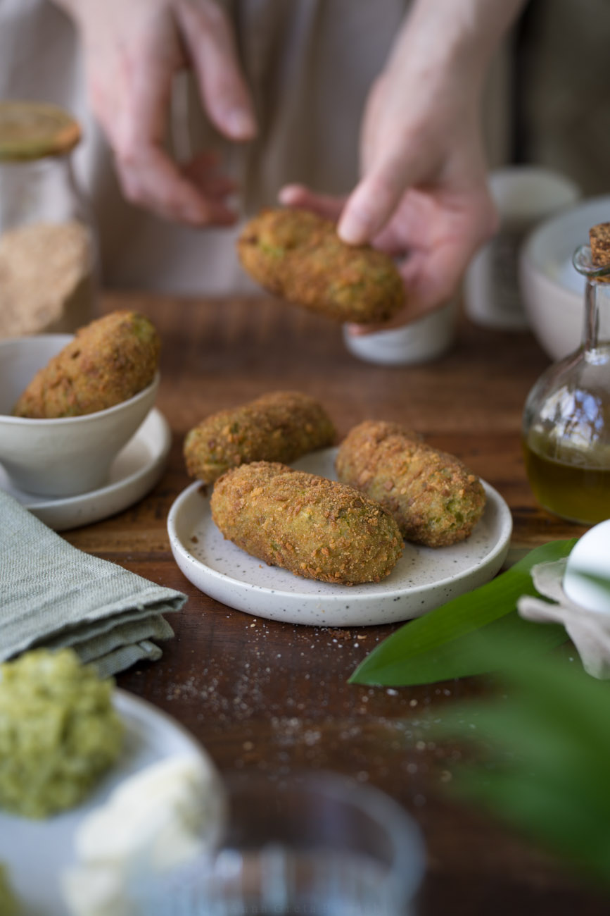 Ein Tellerchen mit drei frittierten Bärlauch-Suppli steht in der Mitte auf einer dunken Holztischplatte. Im Hintergrund sieht man zwei Hände,d ie ein Suppli halten. Vorne sieht man im Anschnitt frischen Bärlauch.