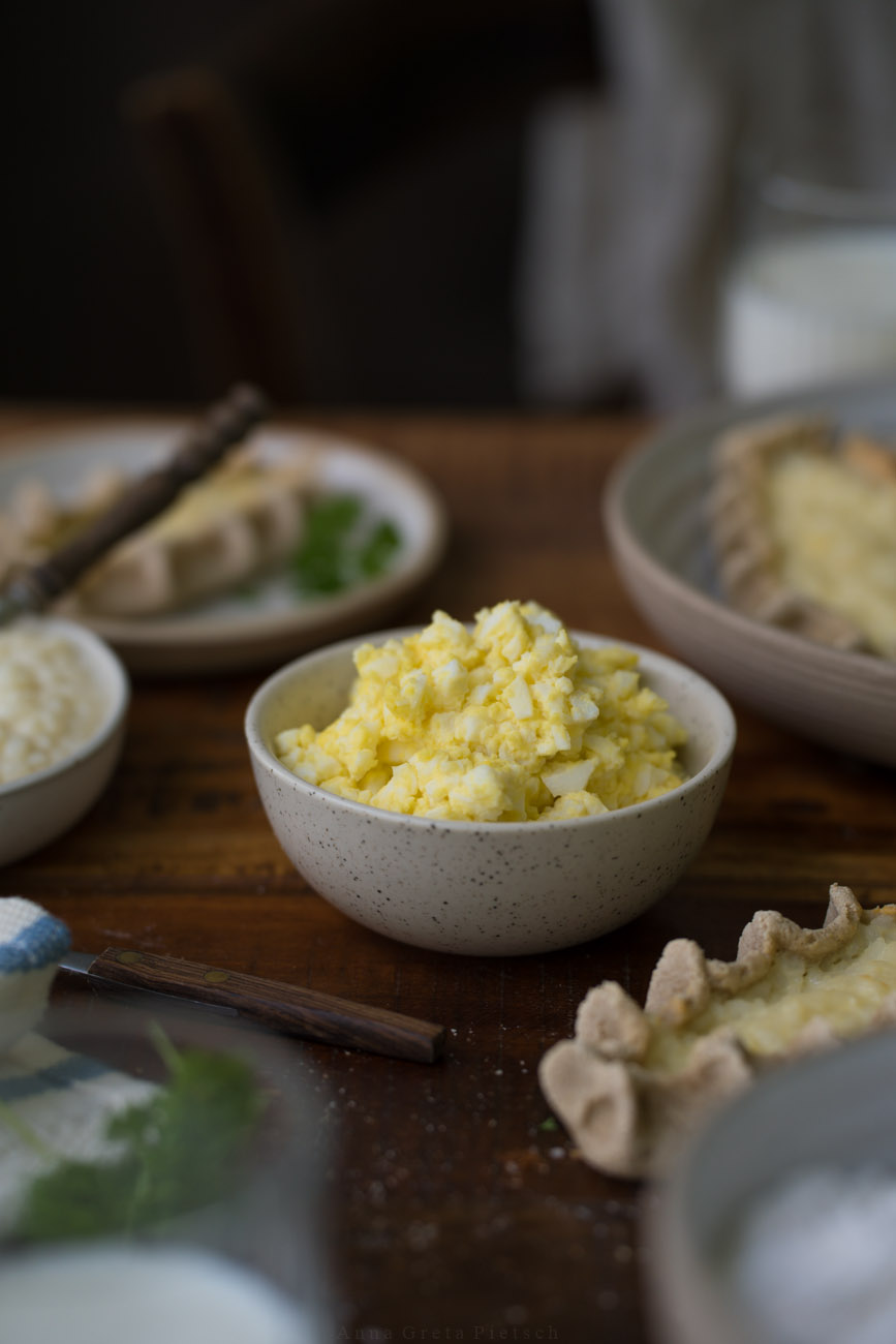 Eine dunkle Holztischplatte, gedeckt mit hellen Tellern. Im Fokus steht eine Schale mit Eibutter (zermengte Eier mit Butter).