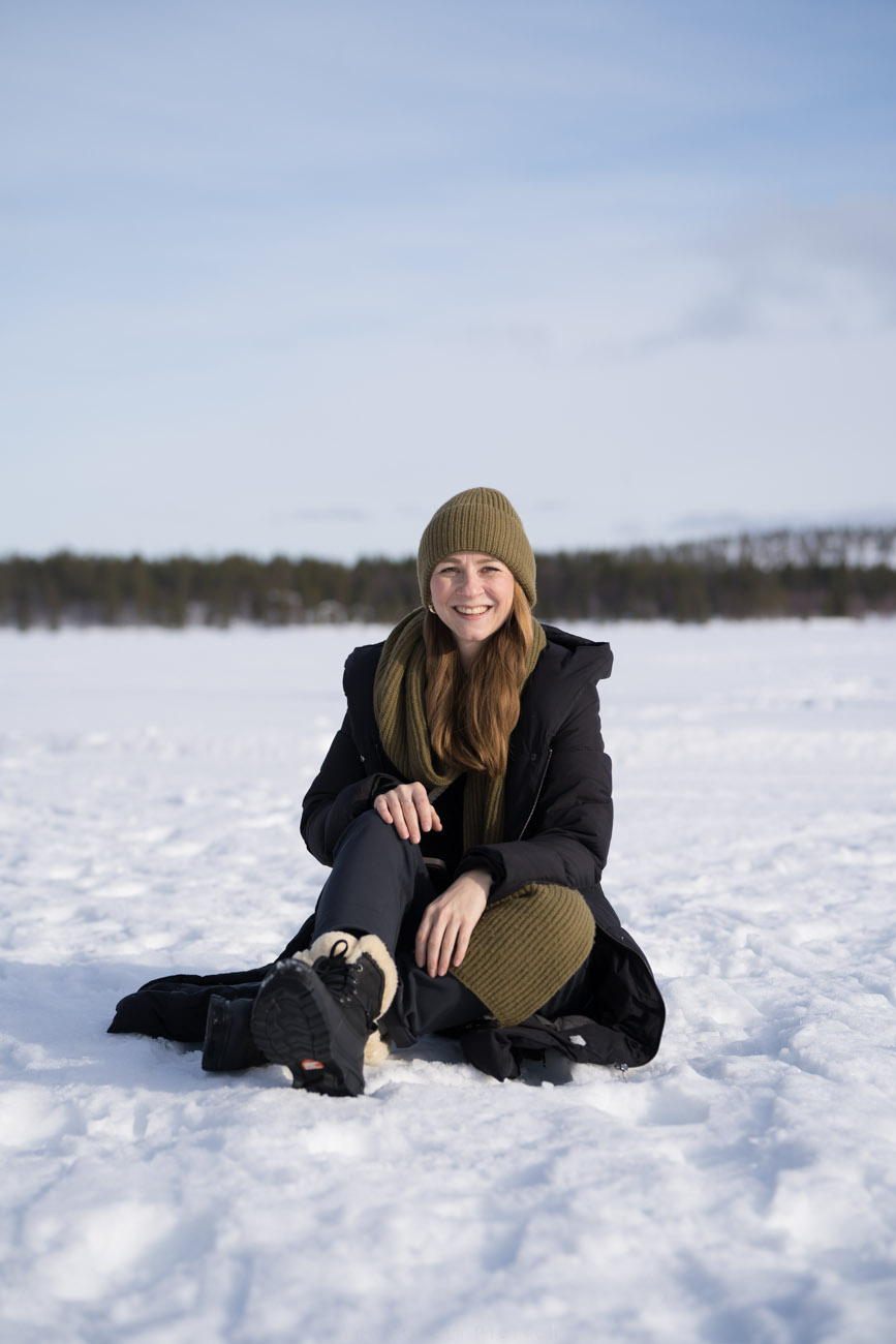 Eine Person mit dicker Winterkleidung sitzt im Schnee auf einem zugefrorenen See und lächelt in die KAmera. Sie trägt eine olivfarbene Mütze und einen dazu passenden Schal und schwarze Kleidung.