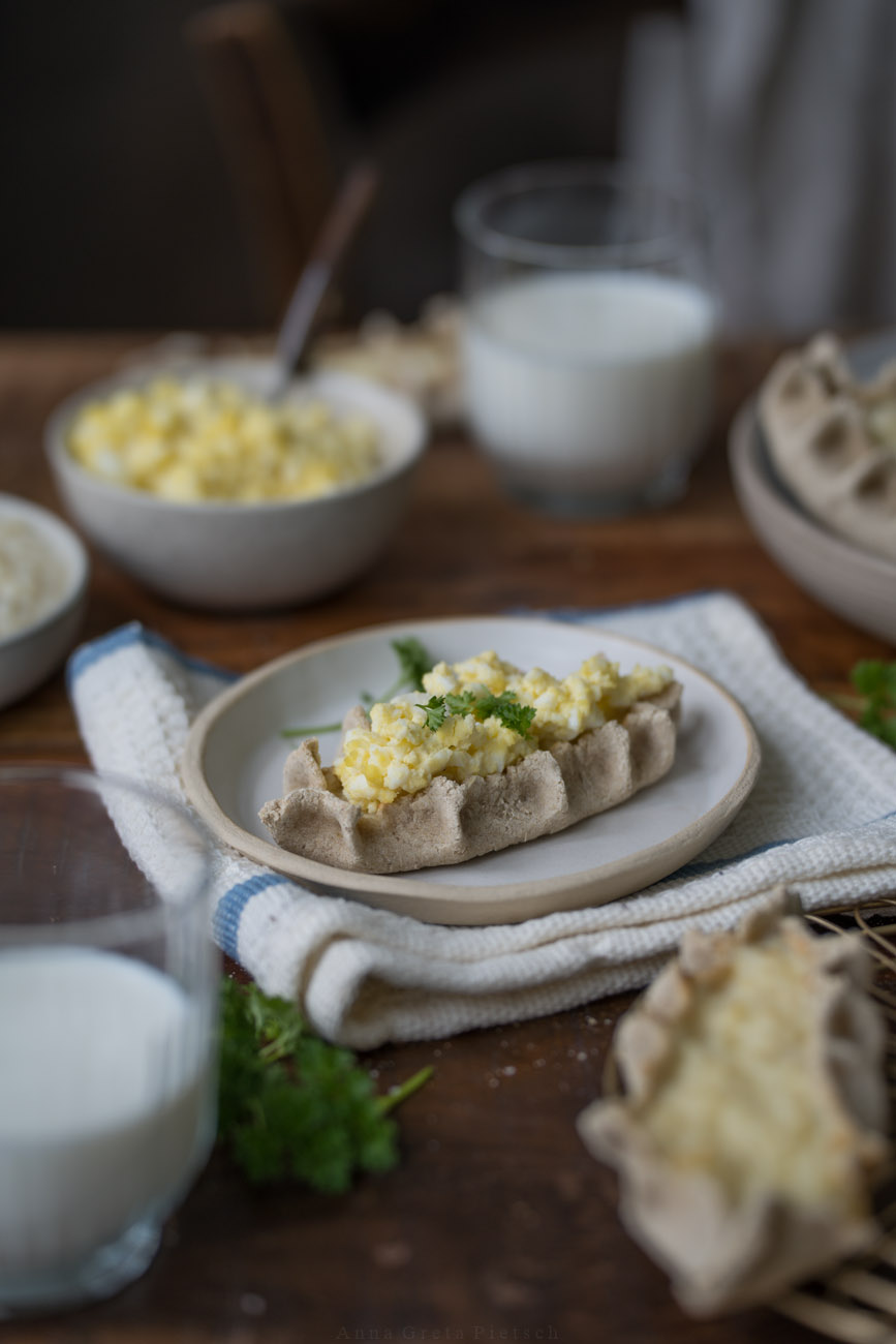Eine dunkle Holztischplatte, gedeckt mit hellen Tellern. Darauf sind finnische Piroggen mit Reisfüllung. 