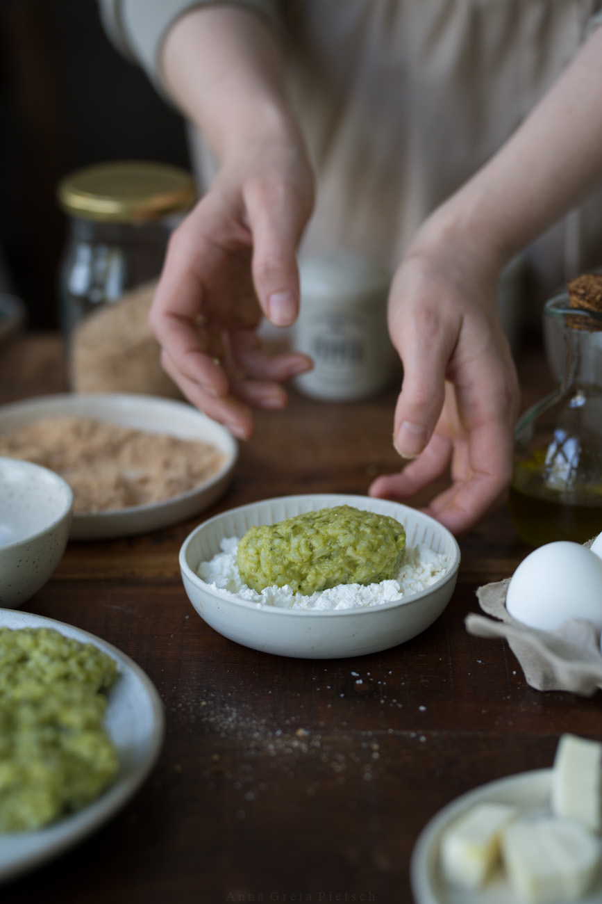Eine helle kleine Keramikschale steht in der Mitte eines dunken Tisches. Darin ist Mehl und eine rohe Reis-Krokette. Zwei Hände sieht man im Hintergrund, die diese gerade panieren. MAn sieht och weitere Teller mit Zuteten darum herumstehen.