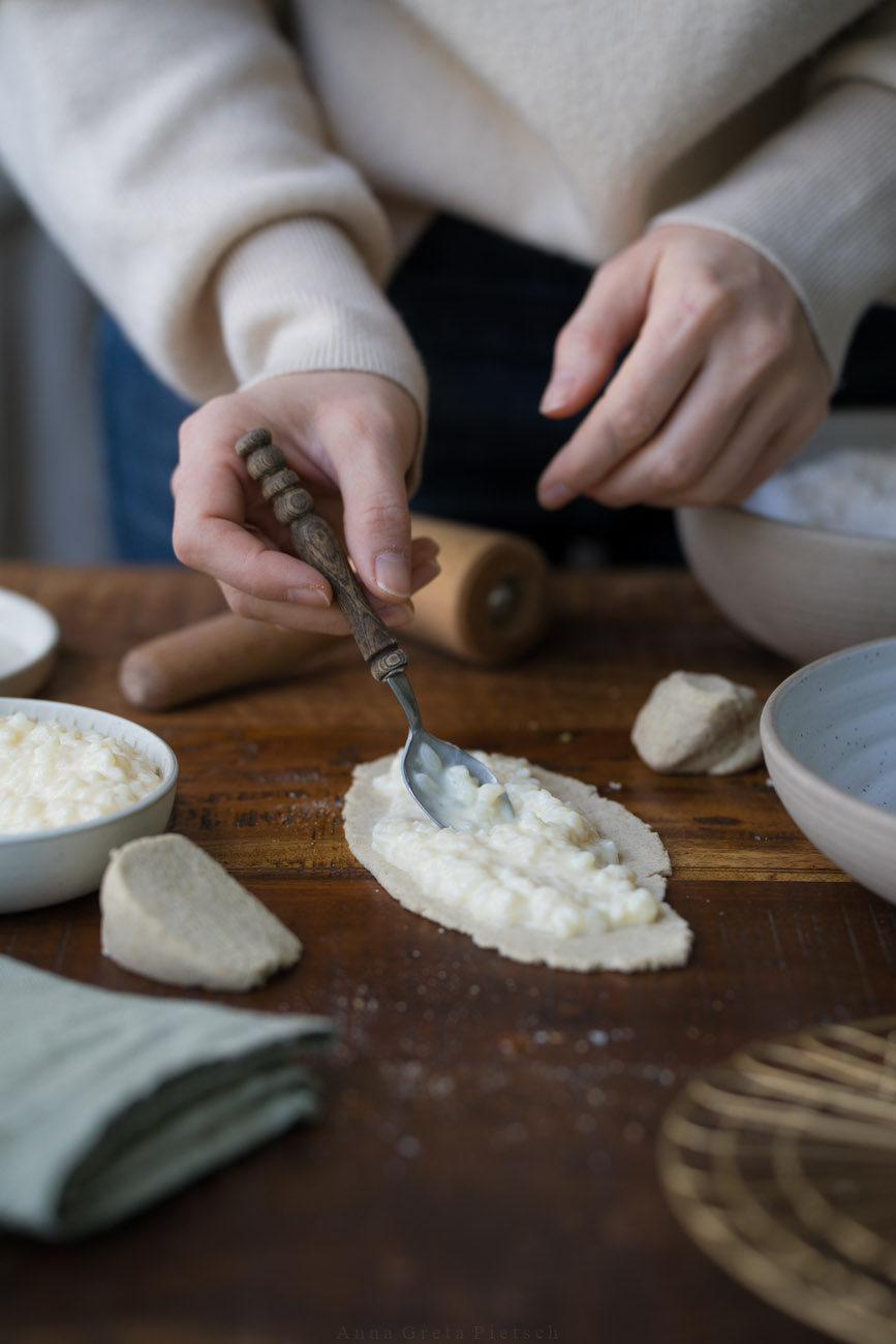 Eine dunkle Holztischplatte, gedeckt mit hellen Tellern. Darauf liegt ein Stückchen ausgerollter Teig und eine Person gibt gekochten Milchreis darauf. So werden Riisipiirakka zubereitet, finnische Piroggen.