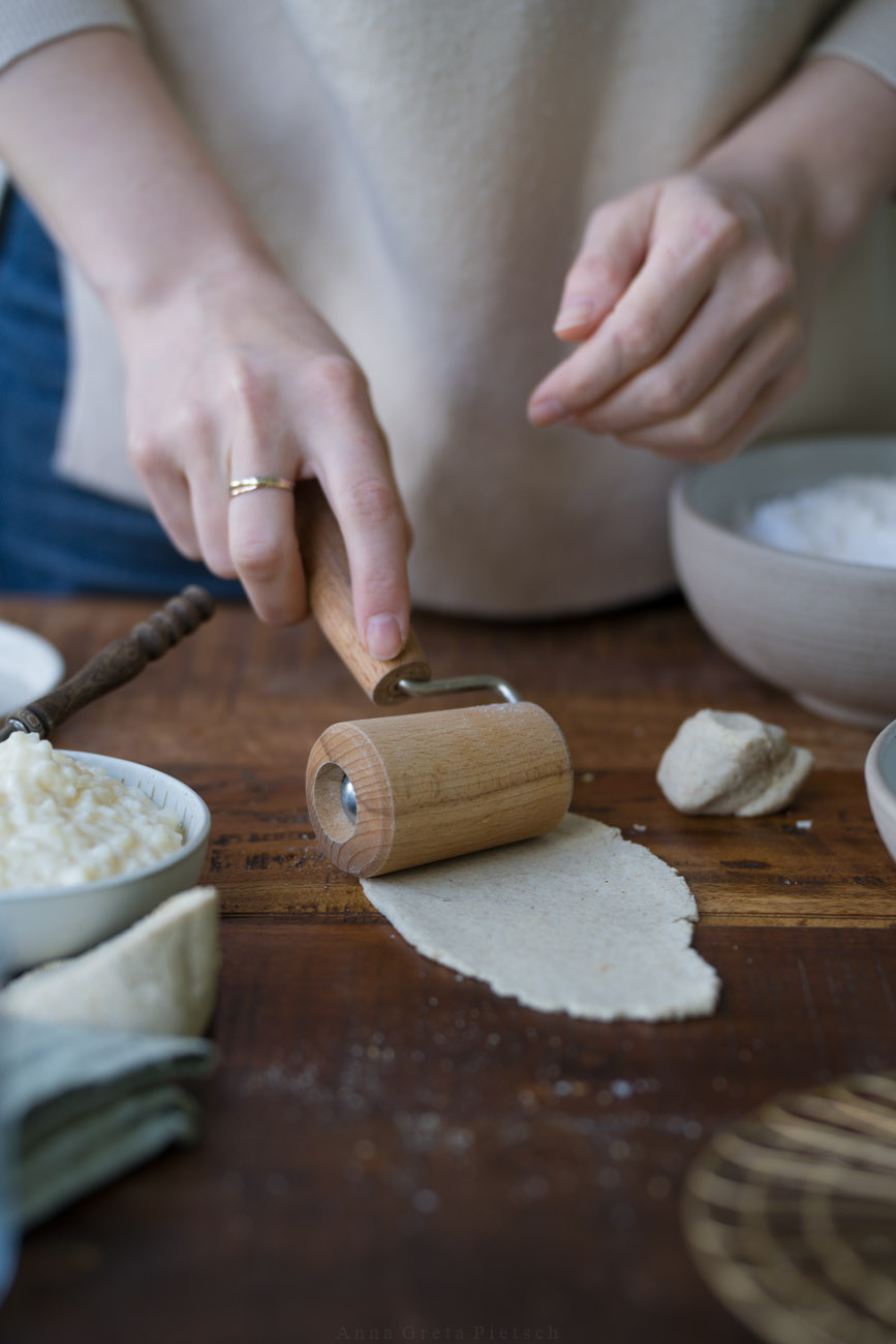 Eine dunkle Holztischplatte, gedeckt mit hellen Tellern. Darauf rollt eine Person gerade ein kleines Stück Teig aus, um die Piroggen zuzubereiten.