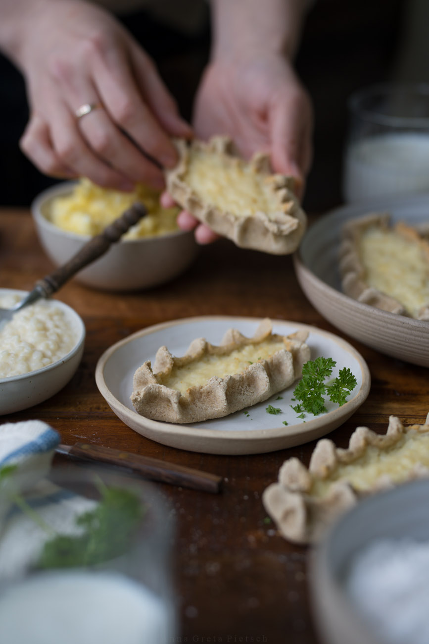 Eine dunkle Holztischplatte, gedeckt mit hellen Tellern. Darauf sind finnische Piroggen mit Reisfüllung. Eine PErson hält eine Pirogge im Hintergrund.