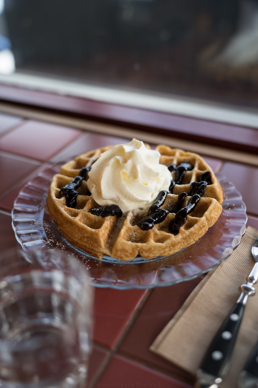 Eine runde Waffel mit Blaubeermarmelade und Sahne auf einem Glasteller. 