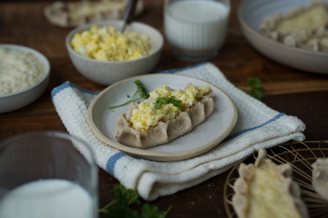 Eine dunkle Holztischplatte, gedeckt mit hellen Tellern. Darauf sind finnische Piroggen mit Reisfüllung.