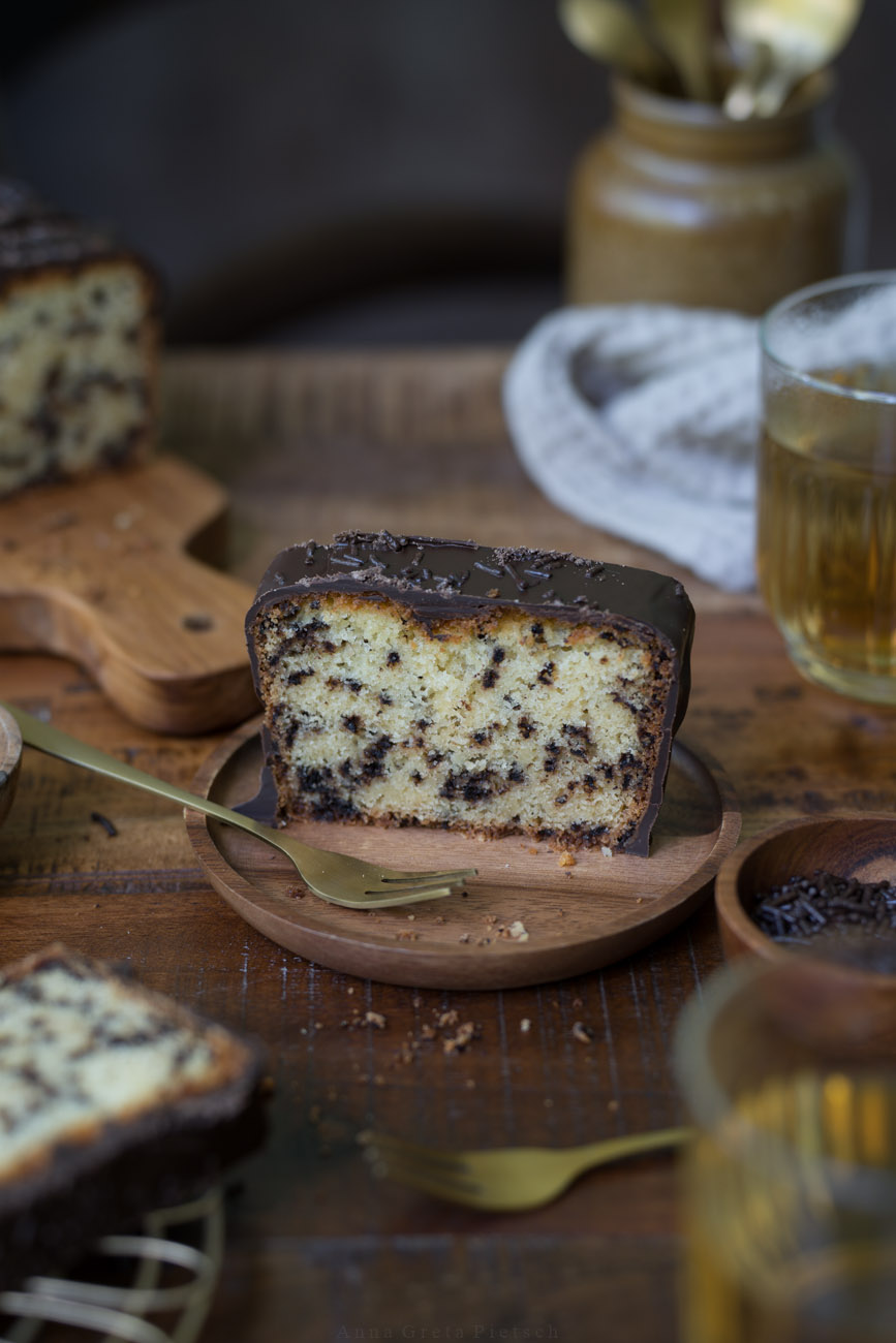 Ein Stück Ameisenkuchen steht auf einem Holztellerchen auf einem Holztisch. Der Tisch ist in natürlichen Farben gedeckt  Man sieht den Kuchenanscnitt im Hintergrund und Teegläser.