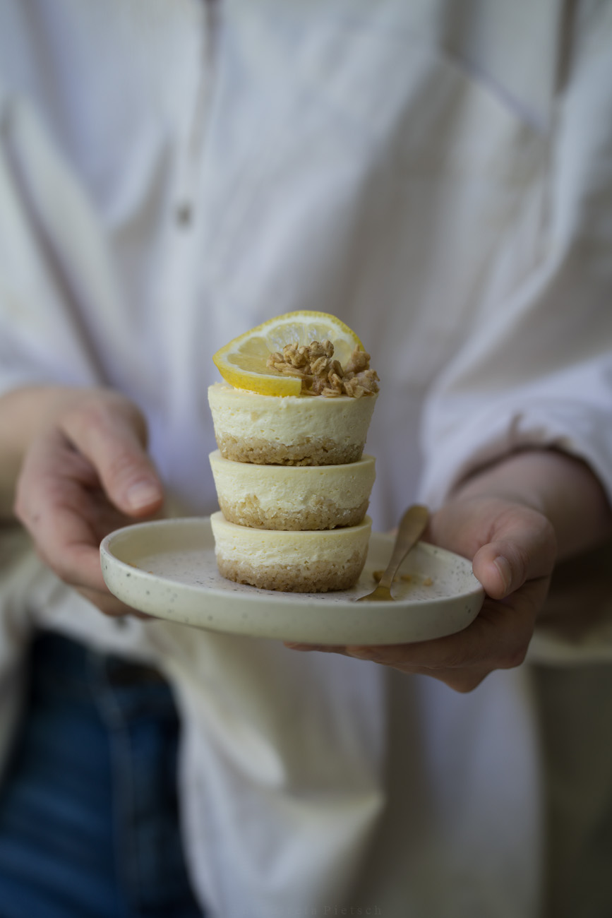 Eine Person mit einer beigen Bluse hält einen kleinen Teller mit drei gestapelten Mini-Cheesecakes in der Hand.