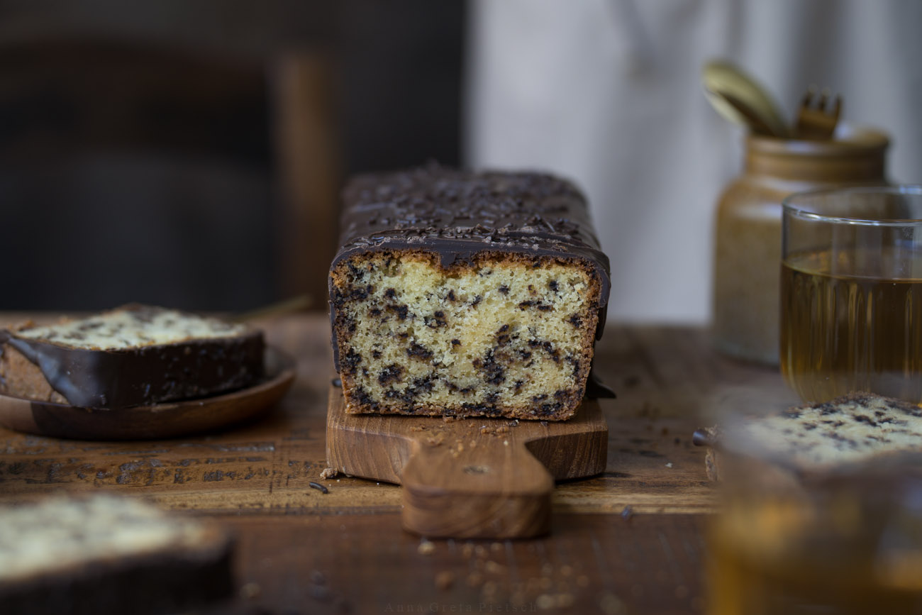Ein Stück Ameisenkuchen steht auf einem Holztellerchen auf einem Holztisch. Der Tisch ist in natürlichen Farben gedeckt Man sieht den Kuchenanschnitt im Hintergrund und Teegläser.