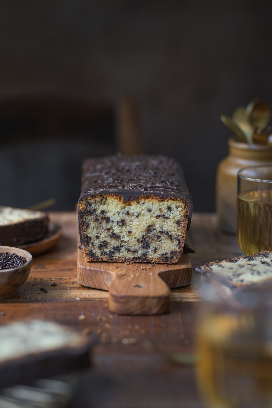 Ein Stück Ameisenkuchen steht auf einem Holztellerchen auf einem Holztisch. Der Tisch ist in natürlichen Farben gedeckt Man sieht den Kuchenanscnitt im Hintergrund und Teegläser.