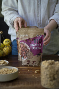Eine Person mit einer beigen Bluse steht hinter einer dunklen Holztischplatte und hält eine Verpackung mit Bio Granola Schoko, die auf dem Tisch steht. Man sieht im Abschnitt noch Schalen und ein Vorratsglas mit Granola.