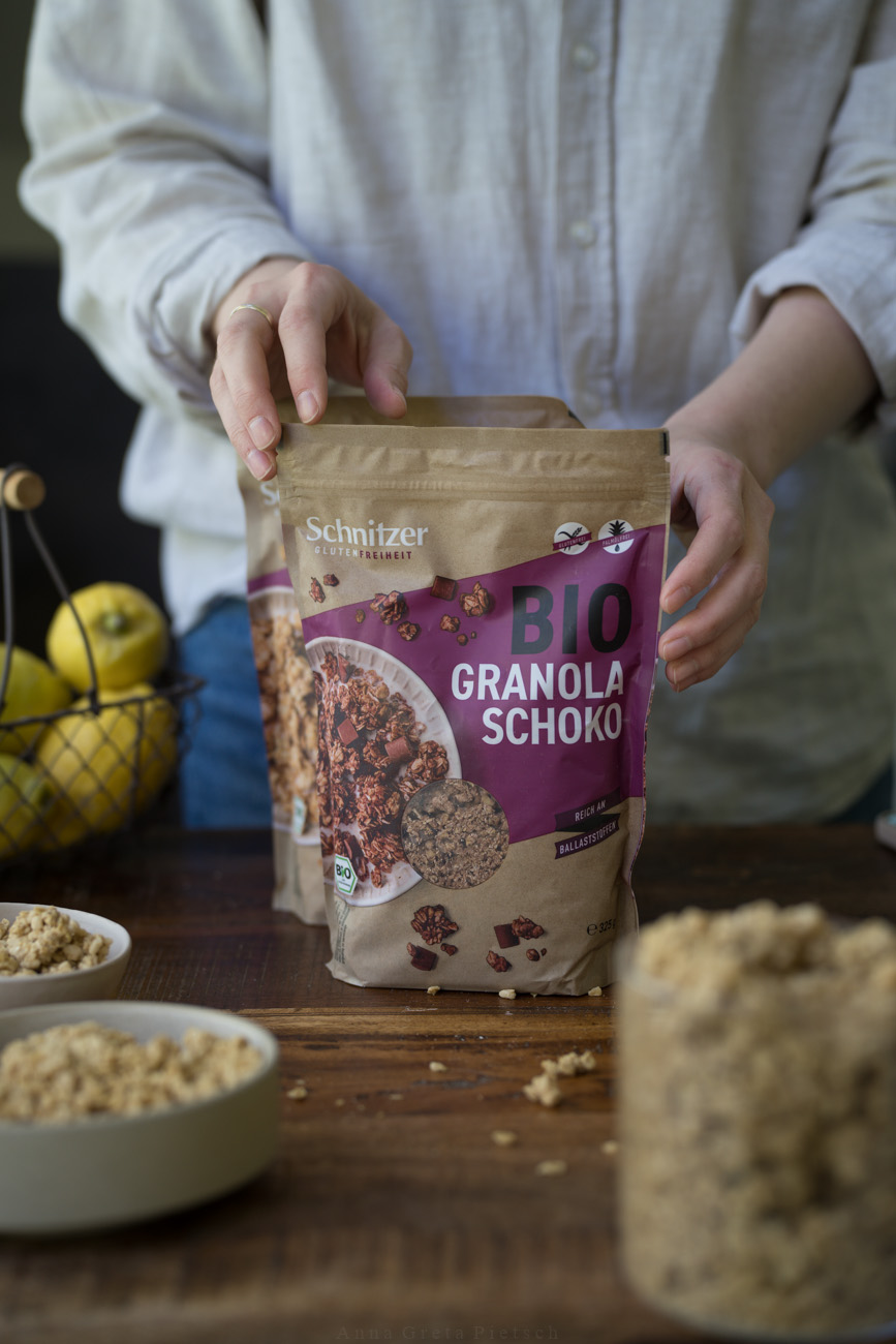 Eine Person mit einer beigen Bluse steht hinter einer dunklen Holztischplatte und hält eine Verpackung mit Bio Granola Schoko, die auf dem Tisch steht. Man sieht im Abschnitt noch Schalen und ein Vorratsglas mit Granola.