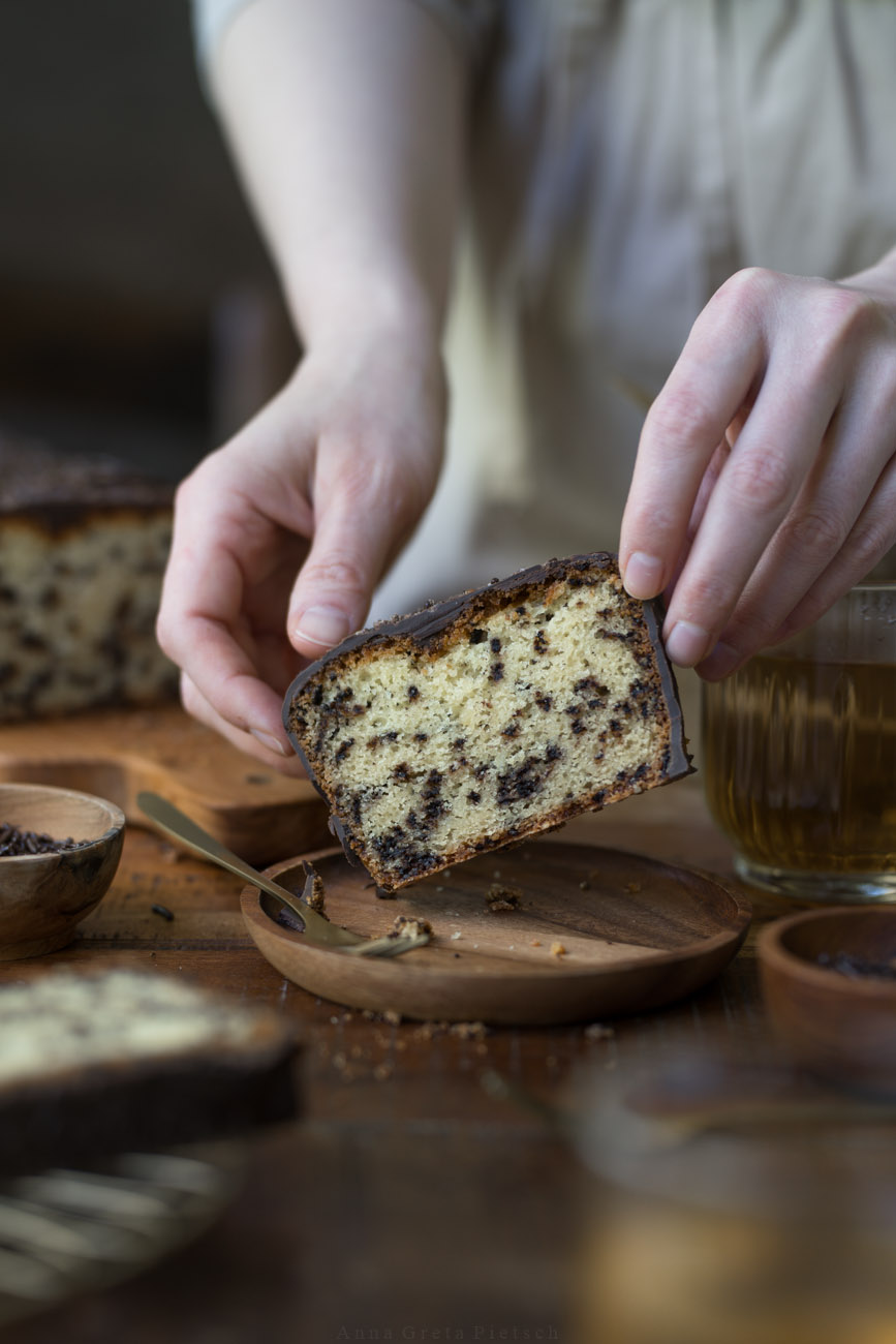 Ein Stück Ameisenkuchen steht auf einem Holztellerchen auf einem Holztisch. Der Tisch ist in natürlichen Farben gedeckt Man sieht den Kuchenanschnitt im Hintergrund und Teegläser.
