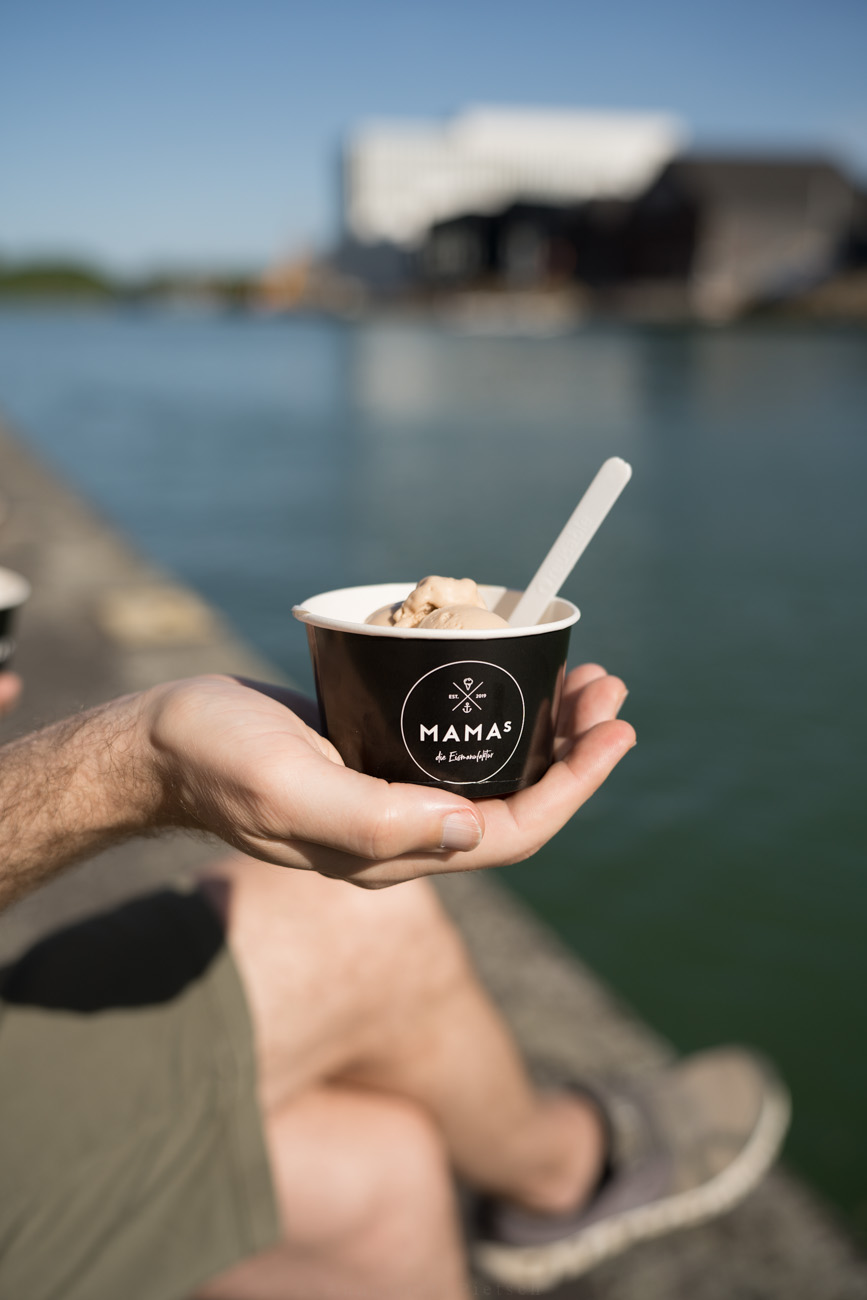 Eien Person hält einen Eisbecher mit einer Kugel eis in der Hand. Im Hintergrund sieht man Wasser und Industriegebäude.