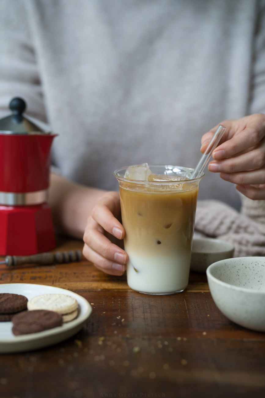 Ein Glas mit einem Eiskaffee steht auf einer Holzplatte. Eine PErson hält den Becher und einen Glasstrohhalm darin. Im Hintergrund steht ein Espressokocher.