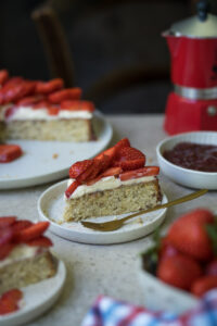 Ein Stück Erdbeerkuchen auf einem kleinen Teller steht in der Mitte einer Kaffeetafel. Man sieht im Hintergrund und Vordergrund noch weitere Kuchenstücke, den Kuchen auf einer Kuchenplatte, Erdbeeren und einen roten Espressokocher.