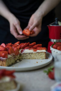 Ein angeschnittener Erdbeerkuchen steht auf einem gedeckten Tisch. Im Hintergrund hält eine Person geschnittene Erdbeerstücke in den Händen.