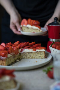 Ein angeschnittener Erdbeerkuchen steht auf einem gedeckten Tisch. Im Hintergrund hält eine Person einen Teller mit einem Stück des Kuchens in den Händen.