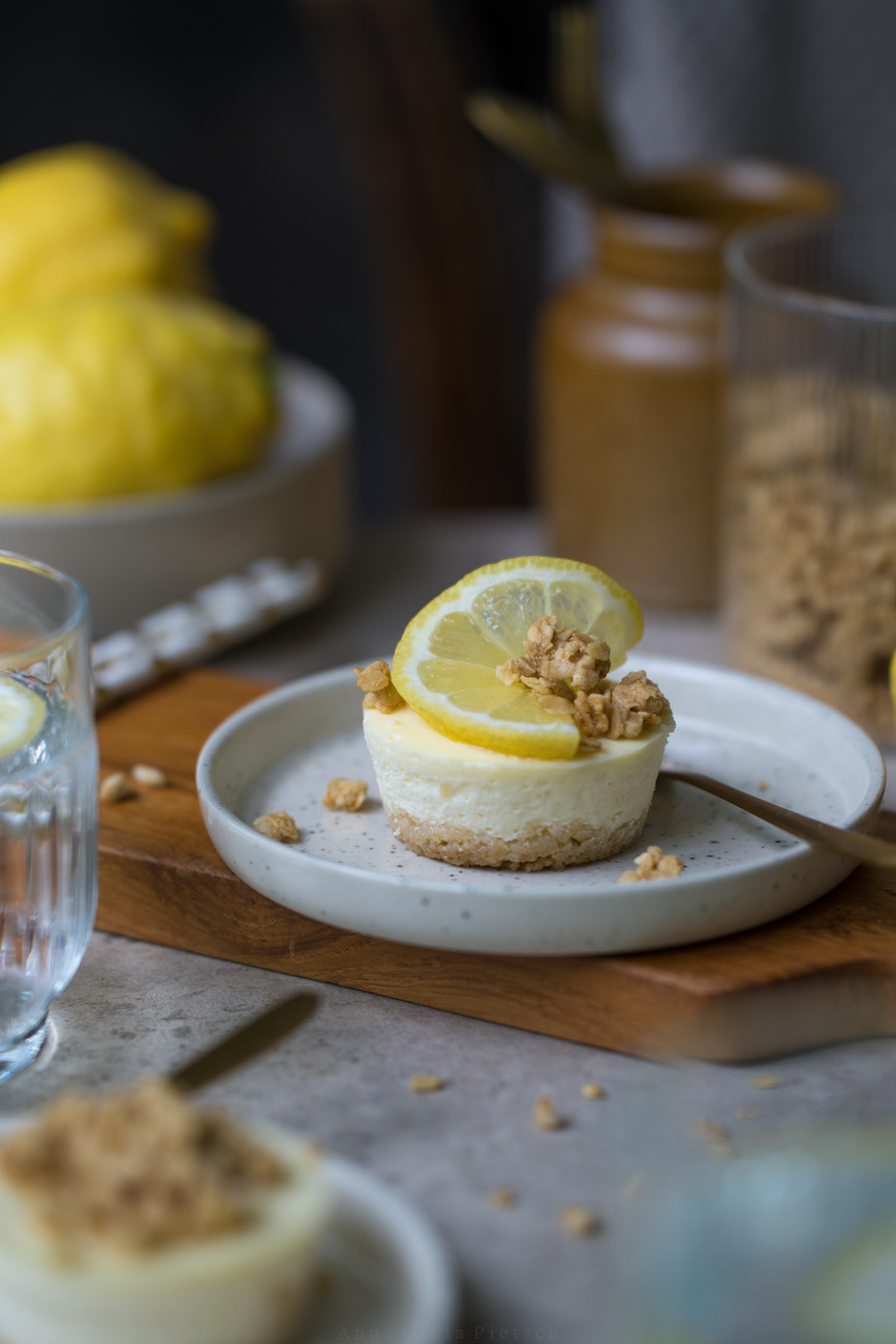 Ein kleiner Teller mit einem Mini-Cheesecakes steht auf einem Tisch. Im Vordergrund sieht man weitere Cheesecakes auf Kuchentellern. Im Hintergrund steht noch eine Schale mit Zitronen.