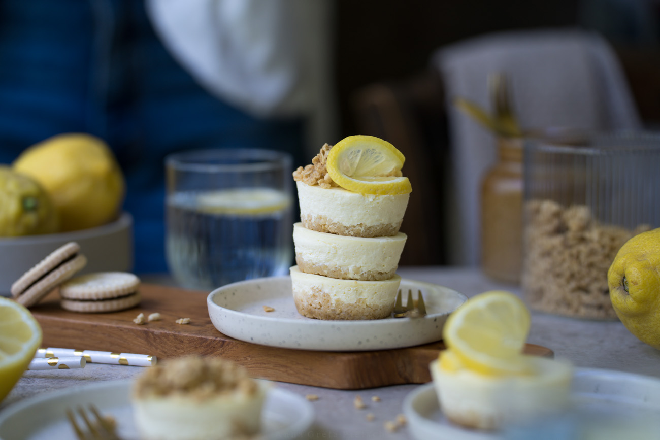 Ein kleiner Teller mit drei gestapelten Mini-Cheesecakes steht auf einem Esstisch. Im Vorder- und Hintergrund sieht man weitere Cheesecakes auf Kuchentellern. Im Hintergrund steht noch eine Schale mit Zitronen.