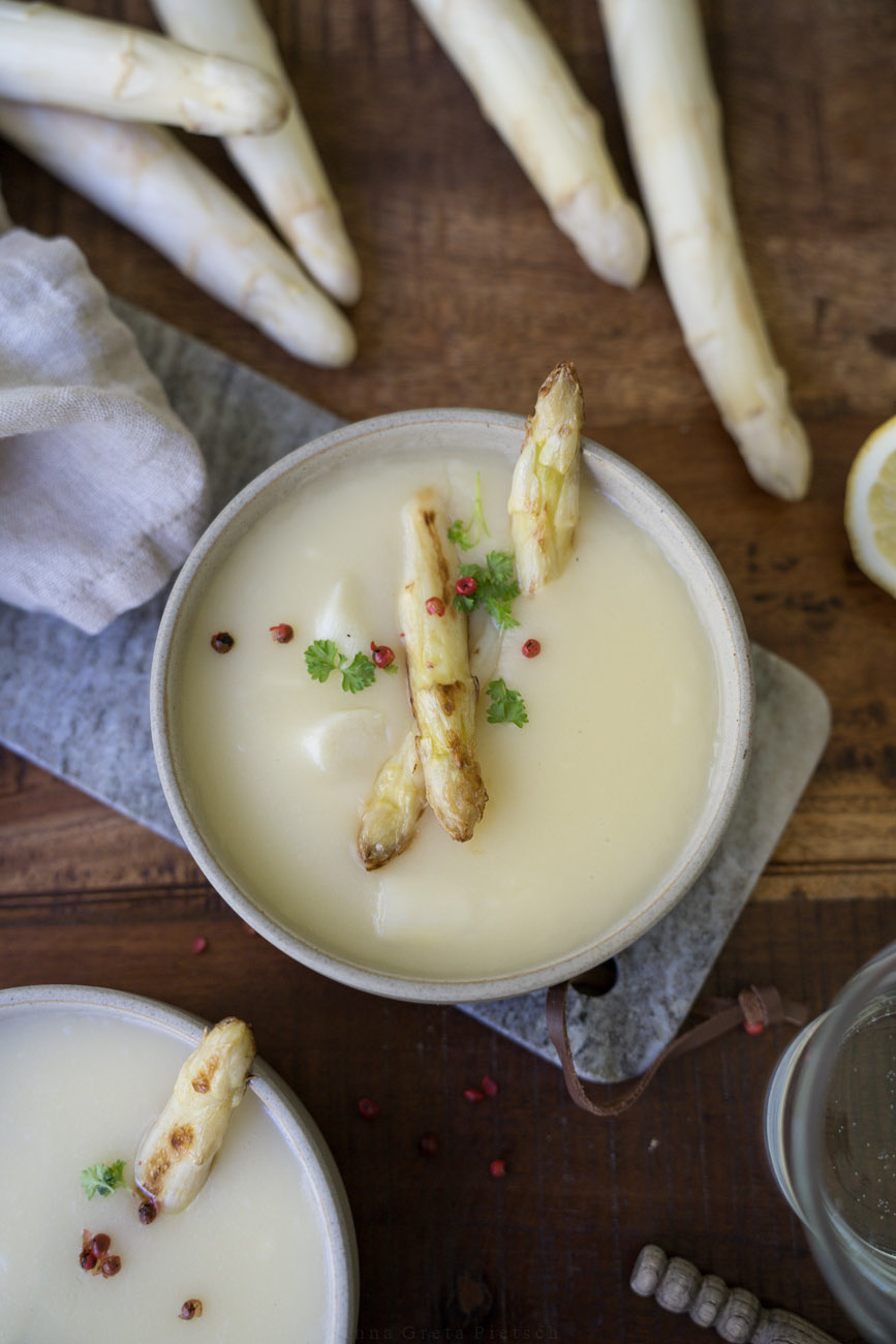 Ein Holztisch, darauf ist Mittagessen angerichtet. In der Mitte steht eine Schale mit Spargelcremesuppe. Im Vordergrund sieht man eine weitere Schale im Anschnitt. Dazu liegen noch Spargelstangen in einer Ecke.