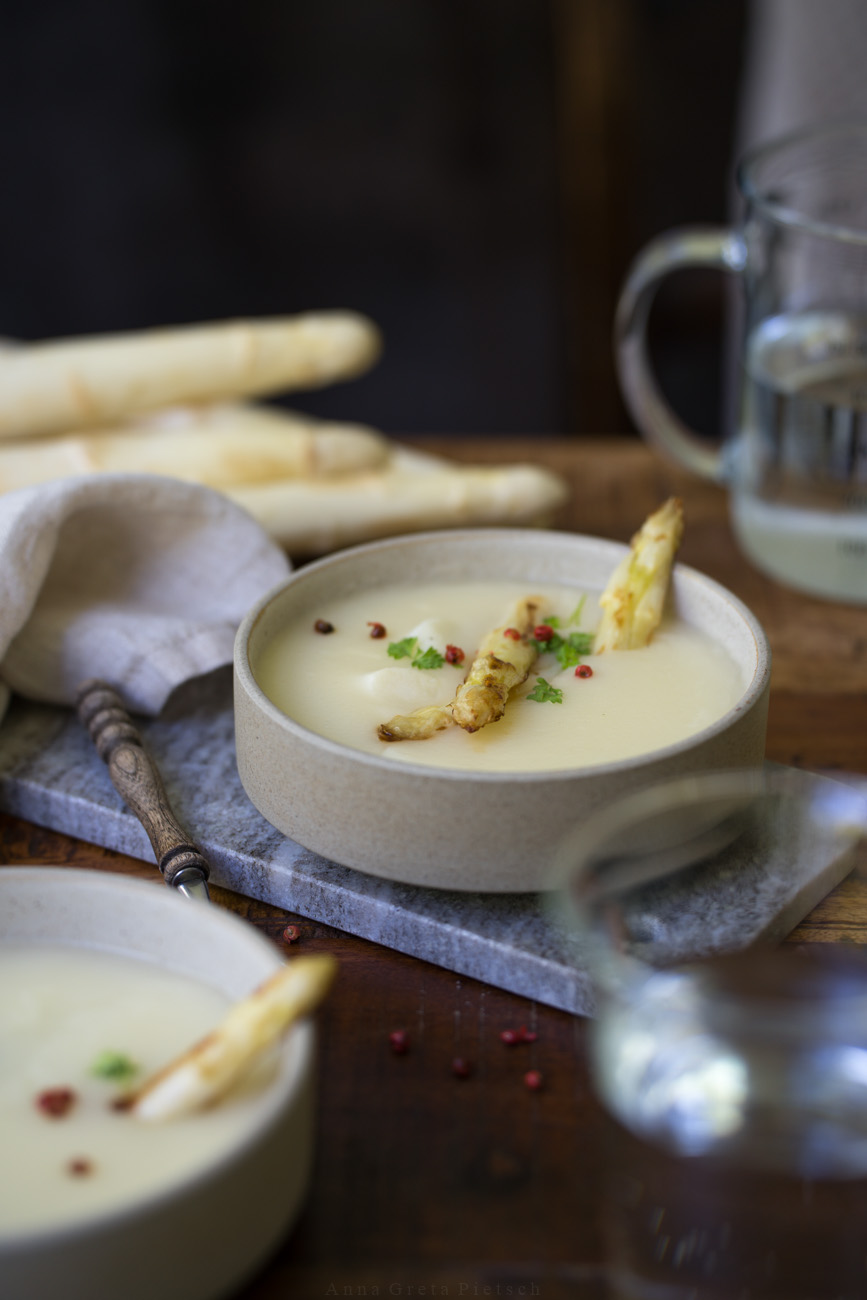 Ein Holztisch, darauf ist Mittagessen angerichtet. In der Mitte steht eine Schale mit Spargelcremesuppe. Im Vordergrund sieht man eien weitere Schale im Anschnitt.