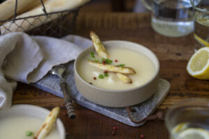 Ein Holztisch, darauf ist Mittagessen angerichtet. In der Mitte steht eine Schale mit Spargelcremesuppe. Im Vordergrund sieht man eien weitere Schale im Anschnitt.