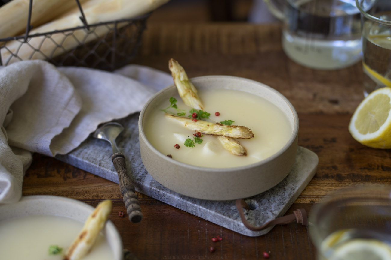 Ein Holztisch, darauf ist Mittagessen angerichtet. In der Mitte steht eine Schale mit Spargelcremesuppe. Im Vordergrund sieht man eien weitere Schale im Anschnitt.