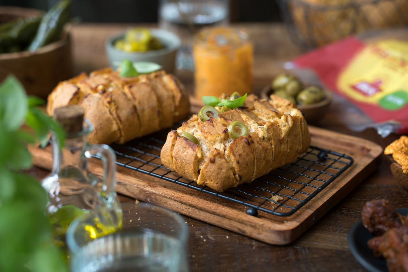 Ein glutenfreies Zupfbrot auf einem Kuchengitter, das auf einer dunklen Holzplatte steht. Drumherum stehen Dips, Oliven und ein Fäschchen mit Öl.