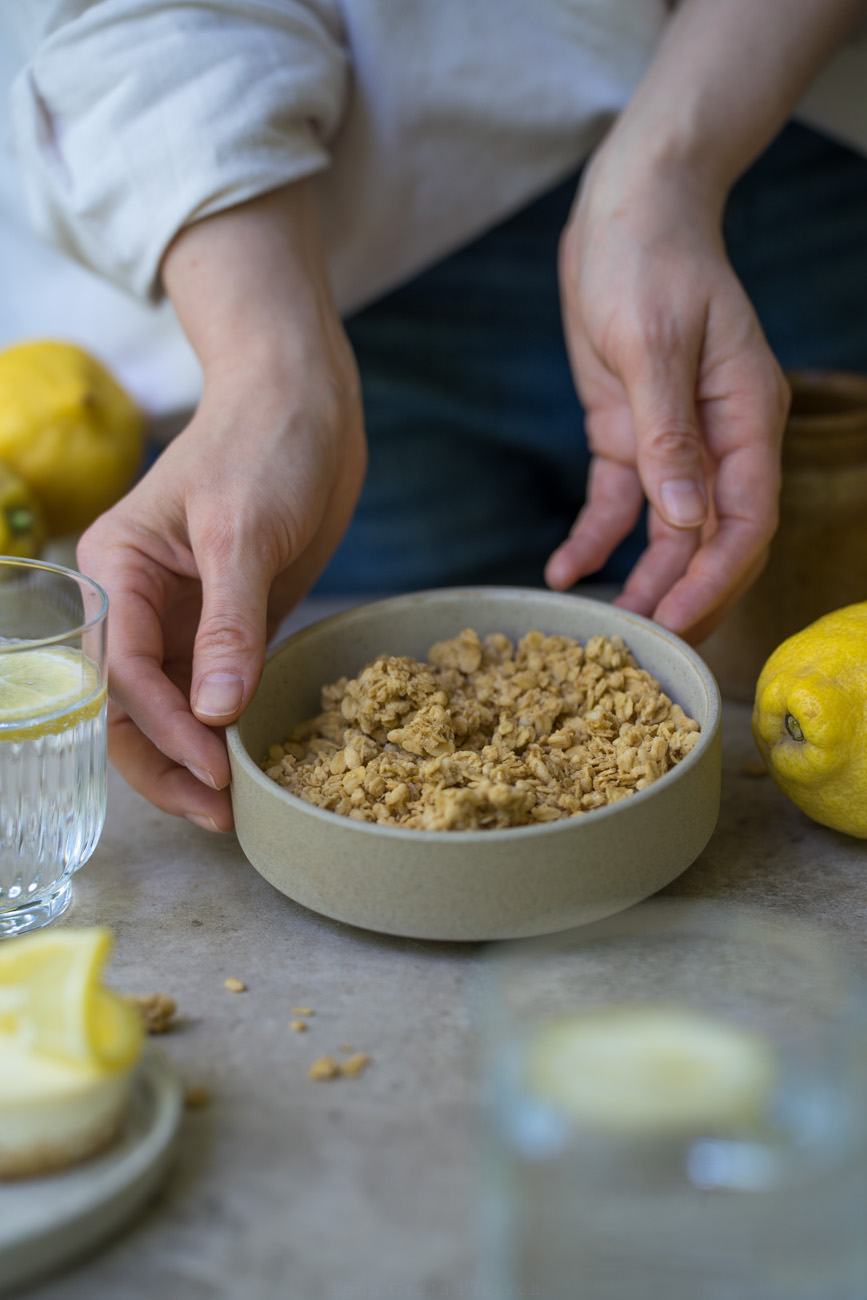 Eine Schale gefüllt mit Bio-Granola steht auf einem dunklen Holztisch. Gehalten wird sie von zwei Händen. Im Hintergrund sieht man ein Vorratsglas mit Granola.