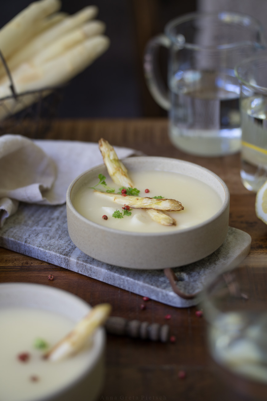 Ein Holztisch, darauf ist Mittagessen angerichtet. In der Mitte steht eine Schale mit Spargelcremesuppe. Im Vordergrund sieht man eien weitere Schale im Anschnitt.