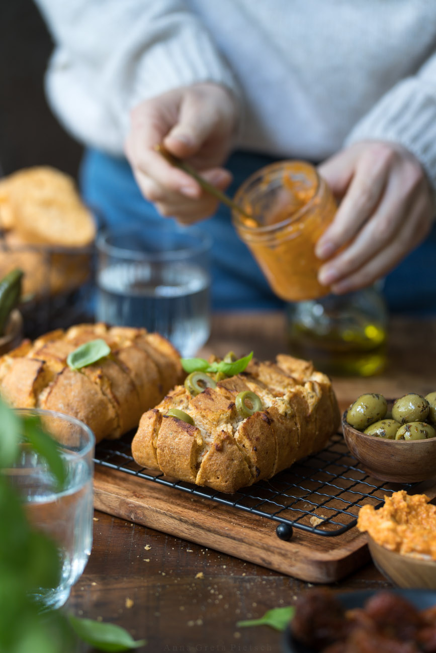 Ein glutenfreies Zupfbrot auf einem Kuchengitter, das auf einer dunklen Holzplatte steht. Drumherum stehen Dips, Oliven und ein Fäschchen mit Öl.