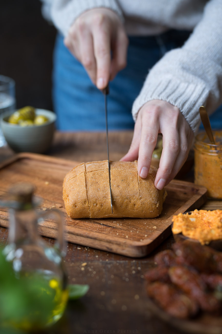 Eine Person mit einem beigen Pullover schneidet ein großes Ciabatta-Brötchen. Drumherum stehen Dips, Oliven und ein Fäschchen mit Öl.