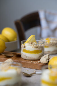 Ein Dessertglas mit Zitronen-Schichtdessert steht auf einem Holzbrettchen auf einer hellen Tischplatte. Im Vorder-/Hintergrund sieht man frische Zitronen und weitere Dessertgläser.