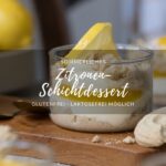 Ein Dessertglas mit Zitronen-Schichtdessert steht auf einem Holzbrettchen auf einer hellen Tischplatte. Im Vorder-/Hintergrund sieht man frische Zitronen und weitere Dessertgläser.