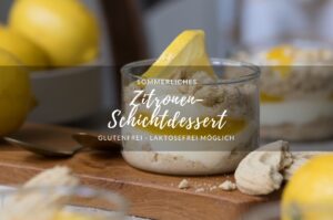 Ein Dessertglas mit Zitronen-Schichtdessert steht auf einem Holzbrettchen auf einer hellen Tischplatte. Im Vorder-/Hintergrund sieht man frische Zitronen und weitere Dessertgläser.
