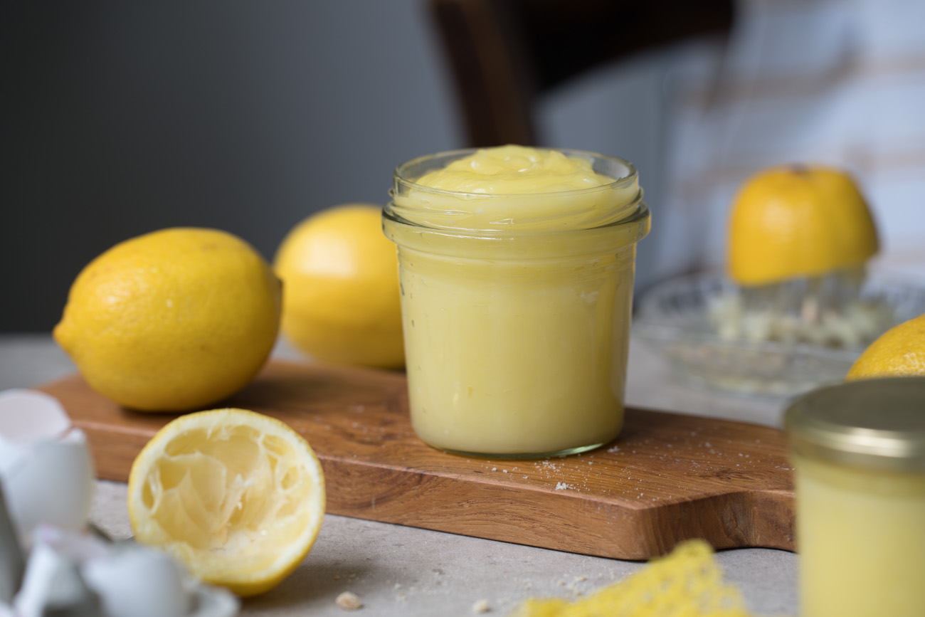 Ein Marmeladenglas mit selbstgemachtem Lemon-Curd steht auf einem Holzbrettchen auf einer hellen Tischplatte. Im Hintergrund sieht man frische Zitronen und im Vordergrund noch ein weiteres gefülltes Glas mit Zitronencreme.