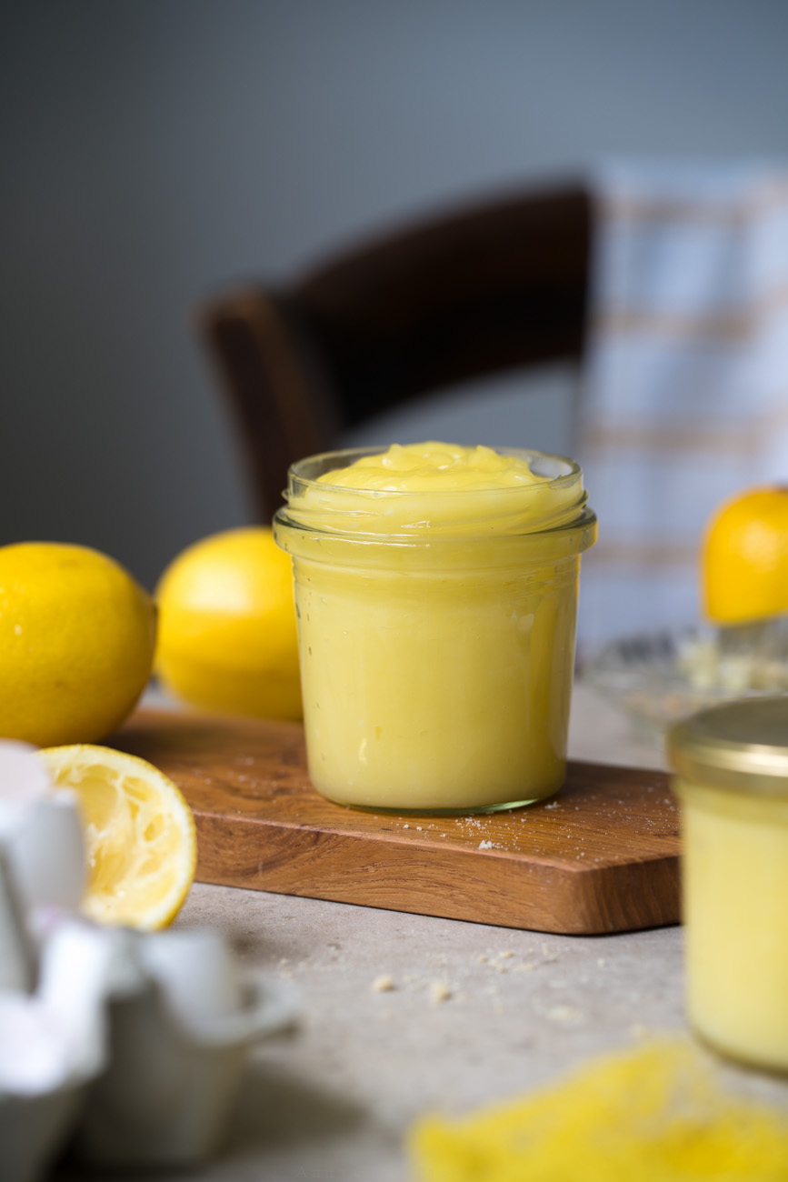 Ein Marmeladenglas mit selbstgemachtem Lemon-Curd steht auf einem Holzbrettchen auf einer hellen Tischplatte. Im Hintergrund sieht man frische Zitronen und im Vordergrund noch ein weiteres gefülltes Glas mit Zitronencreme.