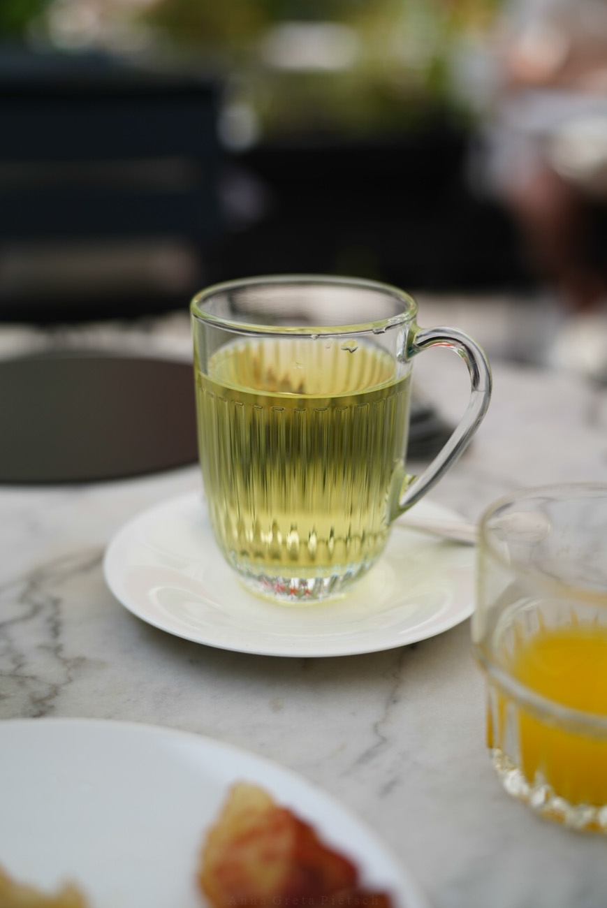 Ein Frühstückstisch auf einer Terrasse mit einem Teebecher aus Glas. Vorne im Anschnitt sieht man ein Glas mit Orangensaft.