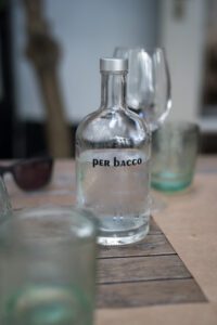 Eine Glasflasche mit Trinkwasser mit der Aufschrift "per bacco" steht auf einem Tisch. Im Vorder- und Hintergrund sieht man Gläser.