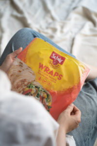 Eine Person hält eine Verpackung mit glutenfreien Wraps von Schär in den Händen/im Schoß. Das Foto wurde über die Schulter aufgenommen.