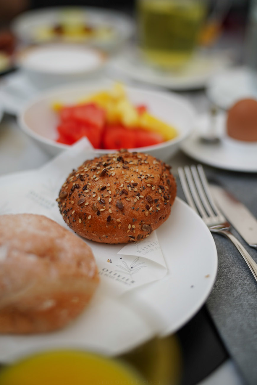 Ein reich gedeckter Frühstückstisch. Im Fokus steht ein rundes Körnerbrötchen. Man sieht weitere Brötchen, Getränke, Obst.