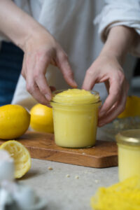 Ein Marmeladenglas mit selbstgemachtem Lemon-Curd steht auf einem Holzbrettchen auf einer hellen Tischplatte. Im Hintergrund sieht man frische Zitronen und im Vordergrund noch ein weiteres gefülltes Glas mit Zitronencreme. Eine Person hält das Glas mit mit beiden Händen fest.
