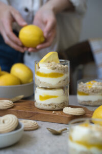 Zwei gestapelte Gläser mit Zitronen-Schichtdessert stehen auf einem Holzbrettchen auf einer hellen Tischplatte. Im Vorder-/Hintergrund sieht man frische Zitronen und weitere Dessertgläser.